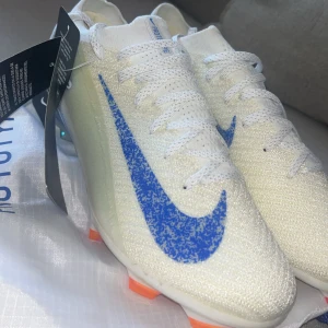 Vita Nike fotbollsskor med blå swoosh - Snygga vita Nike fotbollsskor med en stor blå swoosh på sidan och orange dobbar. Skorna har snörning och är tillverkade i ett lätt syntetmaterial som ger bra känsla på planen. Perfekta för dig som vill sticka ut med stilren design och grym prestanda.