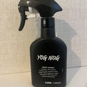 Lush Yog Nog Body Spray 200ml - Yog Nog Body Spray från Lush i svart plastflaska med spraymunstycke. Doften är söt och varm med toner av kola, kryddnejlika och ylang ylang. Flaskan rymmer 200 ml och har en modern, stilren design med vit text på framsidan.