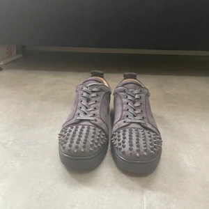 Christian Louboutin svarta sneakers med nitar - Snygga gråa sneakers från Christian Louboutin i mocka med röda sulor och coola nitar på tån. Skorna har snörning och perfekt för dig som vill sticka ut med en edgy vibe. Skorna är i storlek 43 och endast använda 4-5 gånger men sulorna skrapades lite fast kan tas bort om man har rätt medel och därför är priset så billigt.