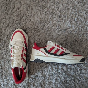 Adidas sneakers i rött, vitt och svart - Snygga Adidas sneakers med klassiska tre ränder i rött och svart på sidorna. Storlek 44.