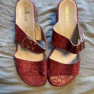 Röda sandaler med korkkilklack från Rohde - Snygga röda sandaler från Rohde med krokopräglat mönster och dubbla remmar med guldfärgade spännen. Skorna har öppen tå och en bekväm korkkilklack som ger extra höjd. Perfekta för sommardagar när du vill ha både stil och komfort.