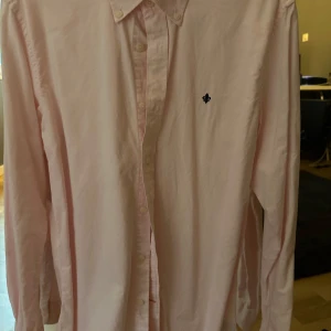 Ljusrosa skjorta från Morris - Snygg ljusrosa skjorta med klassisk krage och knäppning framtill. Skjortan har lång ärm och en liten broderad svart logga på bröstet. Perfekt för dig som gillar stilrena plagg med en twist. Tillverkad i mjuk bomull för skön känsla.