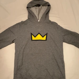 Grå hoodie med gul krona från Delta Fleece - Grå hoodie från Delta Fleece med en stor gul krona tryckt på bröstet. Vet inte storlek men skulle gissa på S