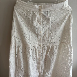 Vit långkjol i broderad bomull Lindex - Superfin vit långkjol från Lindex i broderad bomull med snyggt hålmönster. Kjolen har knappar framtill och volang nedtill för en extra flowy känsla. Perfekt för varma dagar när du vill ha en chill och romantisk look.