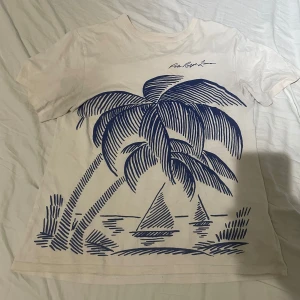 Vit t-shirt Polo Ralph Lauren palmtryck - Vit t-shirt från Polo Ralph Lauren med stort blått palm- och segelbåtsmotiv på framsidan. Klassisk passform med rund hals och korta ärmar. Tillverkad i mjuk bomull, perfekt för sommarens chill dagar. Helt okej skick men fortfarande helt användbar. Storlek 18-20 men passar om man är 175-185