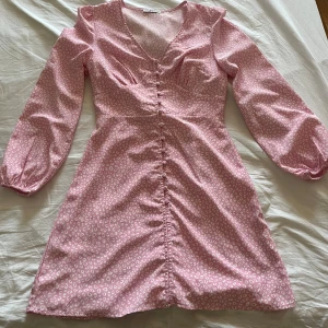 Rosa leopardmönstrad kortklänning - Supersöt rosa kortklänning med vitt leopardmönster och v-ringning. Klänningen har långa ärmar med resår vid handlederna och knappar längs hela framsidan. Det är sytt några extra stygn längst ner på klänningen (se bild) för att täcka, inget som syns speciellt tydligt. Säljes då passformen inte riktigt passade mig🩷