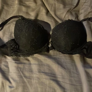 Svart spets-bh med rosett och blommor - Snygg svart bh med vadderade kupor och spetsdetaljer med blommönster. Framtill sitter en liten rosett och en glittrig fjärilsberlock. Axelbanden är justerbara och kanten har fin spets. Perfekt för dig som gillar detaljer och en feminin look.