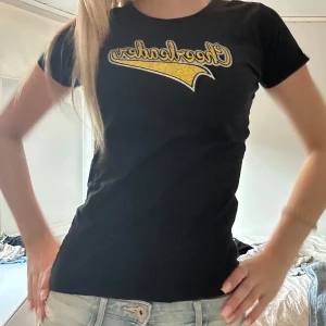 Svart t-shirt med guld - Svart t-shirt med tight passform och korta ärmar. På bröstet finns ett stort tryck i guld och vitt med texten 'Cheerleaders' i en sportig stil. Perfekt för dig som gillar en enkel men ändå cool look.