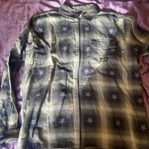 Vailent overshirt med stjärnor L - Snygg overshirt från Vailent i storlek L med dragkedja framtill. Skjortan har rutigt mönster i blått och beige med stjärnor över hela tyget. Bröstficka med etikett och klassisk krage. Perfekt för dig som gillar unika plagg med coola detaljer.