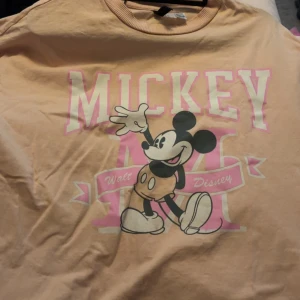 Beige Mickey Mouse t-shirt Disney - Snygg beige t-shirt med stort tryck av Mickey Mouse och texten 'Mickey' i vitt och rosa. Oversized passform och klassisk rund hals. Perfekt för dig som älskar Disney och vill ha en chill och lekfull stil. Storlek M men mer som en L-XL