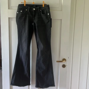 True Religion Joey Low Rise Flare Jeans - Oanvända Snygga gråa jeans från True Religion, modell Joey Low Rise Flare. De har låg midja, utsvängda ben och coola detaljerade bakfickor med silverknappar. Jeansen är i stretchig denim och har en mörk tvätt.