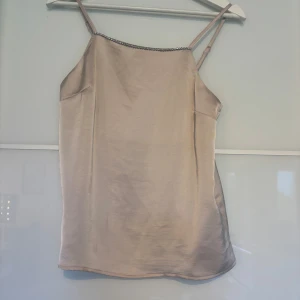 Champagnefärgad linnetopp med strass - Champagnefärgad linnetopp från Kappahl i satinliknande material. Smala axelband och snygg detalj med strass längs halsringningen. Perfekt för dig som gillar en stilren men ändå glammig look. Sitter lätt och luftigt på kroppen.