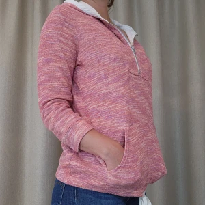 Tommy Hilfiger Rosa hoodie med huva och dragkedja  - Mysig rosa hoodie med vit dragsko och halv dragkedja framtill. Tröjan har en huva med vit insida, känguruficka och ett melerat mönster i rosa och lila toner. Perfekt för chill dagar och enkel att matcha med jeans.