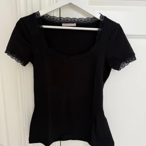 Svart topp med spets  - Svart topp från Anna Field med korta ärmar och snygga spetsdetaljer vid halsringning och ärmslut. Figurnära passform och mjukt material som känns skönt mot huden. Perfekt för dig som gillar en enkel men ändå söt stil.