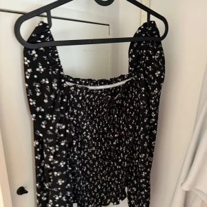 Svart blommig kortklänning med puffärm - Supersöt svart kortklänning med vita små blommor och puffiga långa ärmar. Klänningen har smockad överdel och fyrkantig urringning som ger en trendig vibe. Perfekt för dig som gillar romantiska detaljer och vill sticka ut lite extra.