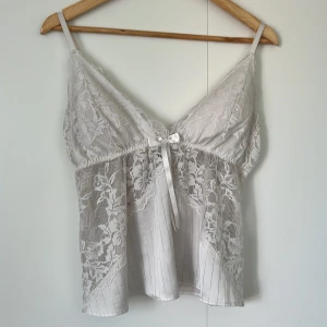 Vit spetsig linnetopp med rosett - Supersöt vit linnetopp med tunna axelband, spetsdetaljer och en liten rosett framtill. Toppen har v-ringning och är tillverkad i ett lätt, glansigt material med blommig spets och diskreta ränder. Perfekt för dig som gillar romantisk och feminin stil.