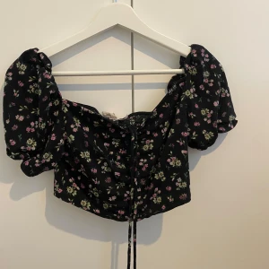 Svart blommig croppad blus H&M S - Supersöt svart croppad blus från H&M med små rosa och gröna blommor. Puffiga korta ärmar och snörning framtill. Går att knyta den kortare