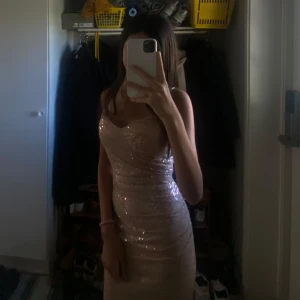 Glittrig beige långklänning - Supersnygg långklänning i beige med glittriga paljetter. Klänningen har smala axelband, figurnära passform och omlott vid bysten. Perfekt för dig som vill sticka ut och glänsa på nästa stora tillfälle!