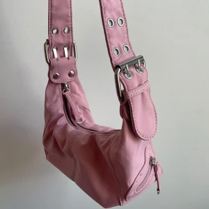 Rosa axelväska med silvriga detaljer - Säljer en söt rosa axelväska med bred axelrem, silvriga metalldetaljer och dragkedja upptill. Väskan har en cool, lite boxig form och extra ficka med dragkedja på sidan. Materialet är mjuk polyester.