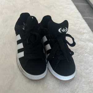 Svarta Adidas Campus sneakers - Svarta Adidas Campus sneakers med klassiska vita ränder på sidorna och vit sula. Skorna har rund tå, snörning och Adidas logga på plös och häl. Ovandelen är i mocka och de har en stilren, tidlös look som passar till det mesta.