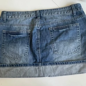 Jeanskjol med nitar och slitningar - Snygg blå jeanskjol med silvriga nitar längs fickorna och slitna detaljer framtill. Kjolen är kort och har klassiska bakfickor midja är 41cm längd är 35cm