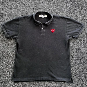 CDG pike - Svart pikétröja från Comme des Garçons Play i bomull med klassisk krage och knäppning framtill. På bröstet sitter det ikoniska röda hjärtat med ögon. Skick 8/10 i använt skick men inga defekter, storlek XL men passar med M/L