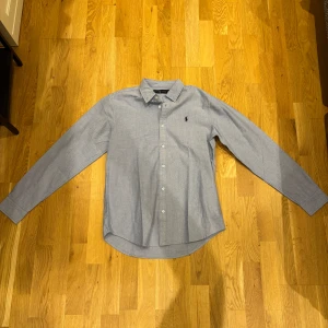 Blå skjorta från Ralph Lauren - Klassisk ljusblå skjorta från Ralph Lauren med button-down krage och broderad logga på bröstet. Skjortan har långa ärmar, knäppning framtill och är tillverkad i mjuk bomull. Perfekt för en clean och stilren look. Ny skick! Nypris runt 1200