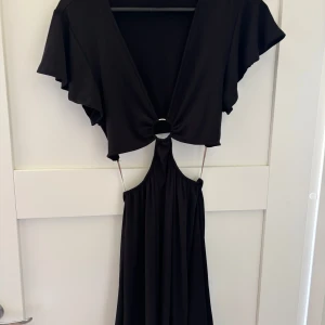 Svart långklänning med ringdetalj - Svart långklänning med djup v-ringning och snygg ringdetalj framtill. Klänningen har vida korta ärmar och öppen midja med smala band. Perfekt för dig som vill sticka ut med en unik och trendig look.