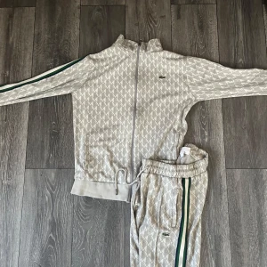 Mönstrade mjukisbyxor från Lacoste - Snygga ljusgrå mjukisbyxor från Lacoste med unikt geometriskt mönster och grönt Lacoste-märke. Byxorna har gröna och vita ränder längs sidorna och resår i midjan för skön passform. Perfekta för en chill och sportig look.
