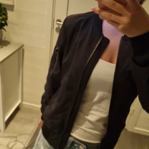 Svart bomberjacka med camofoder - Säljer en svart bomberjacka med klassisk passform och dragkedja framtill. Jackan har en ficka med dragkedja på ärmen och insidan är fodrad med camouflagemönster. Perfekt för dig som vill ha en stilren men ändå edgy look.