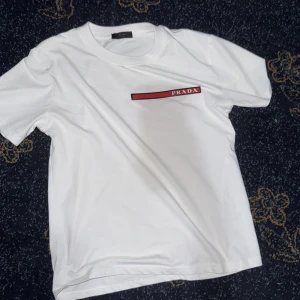 Vit t-shirt från Prada med logga - Snygg vit t-shirt från Prada med klassisk rund hals och korta ärmar. På bröstet finns en röd och svart detalj med PRADA-loggan tryckt i vitt. T-shirten är i mjuk bomull och har en clean, minimalistisk look som passar till allt.