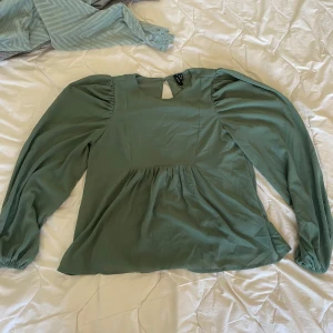Grön blus med puffärm från Vero Moda - Supersöt grön blus från Vero Moda i storlek XS. Blusen har långa puffärmar med resår vid handlederna och en lätt rynkad midja som ger en flowy känsla. Rund halsringning och knapp i nacken för en snygg detalj. Perfekt för dig som gillar stilrena och trendiga plagg.