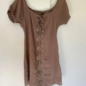 Brun klänning - Miniklänning i ett tunt material, perfekt till sommaren. Nästan en blandning mellan klänning och tunika. På framsidan har den fina paljettdetaljer samt en snörning. Storlek S!