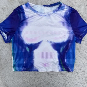 Blå och vit croppad topp med tryck - Cool croppad topp med kort ärm och rund hals. Toppen har ett unikt abstrakt tryck i blått, vitt, lila och grönt med inslag av tunna linjer och färgskiftningar. Materialet är stretchigt och känns syntetiskt, perfekt för en trendig look.