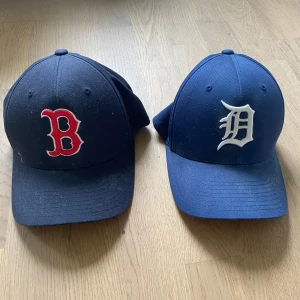 Blåa kepsar Boston Red Sox & Detroit - Två klassiska basebollkepsar i mörkblått, en med Boston Red Sox röd B-logga och en med Detroit Tigers vit D-logga framtill. Båda har böjd skärm och är tillverkade i bomull. Perfekta för sportiga outfits eller streetwear.