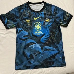 Nike Brasil fotbollströja med pantermotiv - Unik Brasil-fotbollströja från Nike med ett stort blått pantermotiv och gula detaljer. Tröjan har korta ärmar, svart krage och är tillverkad i ett lätt, sportigt material. Brasil-logga och Nike Swoosh i gult framtill. Perfekt för dig som vill sticka ut på planen.