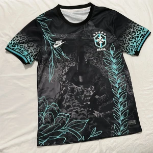 Nike Brasil svart fotbollströja leopard - Unik svart Brasil-fotbollströja från Nike med turkosa detaljer, leopardmönster på ärmarna och stora grafiska blad och blommor. Tryckt Brasil-märke och Nike-logga på bröstet. Tillverkad i lätt och ventilerande material, perfekt för match eller träning.