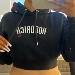 Svart croppad hoodie från Hoodrich - Svart croppad hoodie från Hoodrich med vit broderad logga framtill och coola broderade detaljer på huvan i vitt och rött. Tröjan har snörning i huvan och bred mudd nedtill för en snygg passform. Perfekt för dig som gillar streetwear vibes.