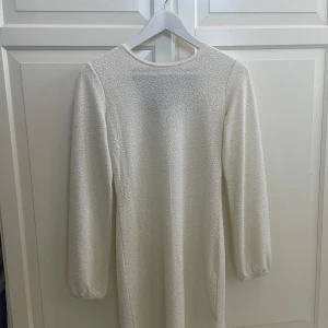 Glittrig vit klänning med rosett Lindex - Supersöt vit glittrig klänning från Lindex med lång ärm och svart rosett i ryggen. Klänningen har en lätt skimrande yta och rund halsringning. Perfekt för jul, eller nyår. Aldrig använd 