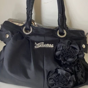 Svart handväska med blommor från Guess - Svart handväska från Guess med coola blommor framtill och silvriga detaljer. Väskan har två handtag, dragkedja upptill och ett mönstrat innerfoder i beige och grått. Insidan har flera fack och Guess-logga i metall. Perfekt för dig som gillar statement-accessoarer.