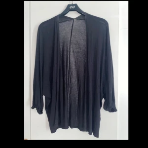 Svart loose kofta i tunn bomull - En svart, tunn och flowy kofta med loose passform och långa ärmar. Perfekt att slänga över en t-shirt eller linne för en chill och avslappnad look. Materialet känns lätt och svalt, vilket gör den enkel att styla året runt.