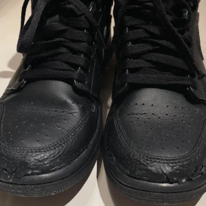 Svarta Nike Air Jordan 1 Mid - Säljer ett par helsvarta Nike Air Jordan 1 Mid med klassisk siluett och snörning. Skorna är i skinn med perforerade detaljer på tån och har en rund tå samt platt sula. Perfekta för dig som gillar en clean och tidlös look.