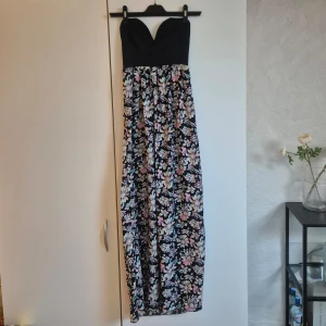 Blommig långklänning från ICE Fashion - Långklänning från ICE Fashion, Australien. Med svart axelbandslös överdel som är smickrande för bysten och en flowy kjol med stort blommönster i pastellfärger. Perfekt för sommarens alla tillfällen! Söt sweetheart-ringning och mjukt, lätt material.