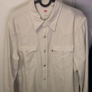 skjorta från Levi's - Snygg vit westerninspirerad skjorta från Levi's med klassisk spetsig krage, tryckknappar och två bröstfickor med lock. Skjortan är i bomull och har långa ärmar samt coola sömdetaljer på axlar och rygg. Perfekt för dig som gillar stilren och tidlös look.