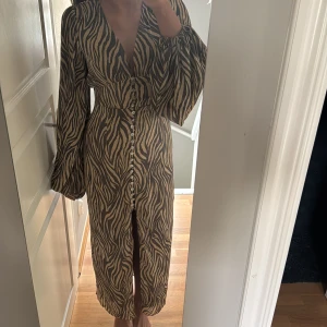 Zebramönstrad långklänning med knappar - Snygg långklänning med zebramönster i beige och svart. Djup v-ringning, markerad midja och långa puffärmar. Klänningen har en rad pärlemorknappar framtill och är tillverkad i ett lätt, flowy material. Perfekt för dig som vill sticka ut med en cool look. Saknas en knapp längst nere men det är inget men tänker på, då jag tycker de blev fint ☺️