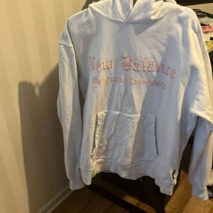 Vit hoodie New Balance med skelettprint - Vit hoodie från New Balance med rosa broderad text framtill och stor svart skelettillustration på ryggen. Klassisk känguruficka, huva och lång ärm. Perfekt för dig som gillar streetwear och unika detaljer.