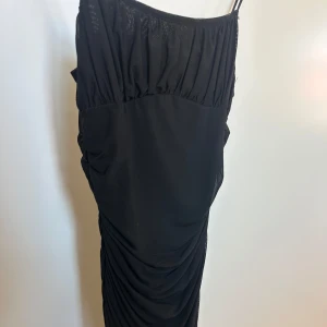 Svart klänning från SHEIN - Svart klänning från SHEIN med smala axelband och rynkade detaljer längs sidorna. Klänningen har en figurnära passform och är tillverkad i ett lätt, stretchigt syntetmaterial. Perfekt för dig som gillar en stilren och trendig look.