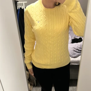 Gul kabelstickad tröja från GANT - Snygg gul kabelstickad tröja från GANT med rund halsringning och broderad logga på bröstet. Tröjan är i bomull och har ribbade muddar vid ärmslut och nederkant. Perfekt för dig som gillar preppy och färgglada vibes.