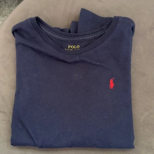 Mörkblå långärmad tröja Polo Ralph Lauren - Mörkblå långärmad tröja från Polo Ralph Lauren med klassisk röd broderad logga på bröstet. Tröjan är i mjuk bomull och har en enkel, stilren design med rund hals. Perfekt för dig som gillar en clean och tidlös look.