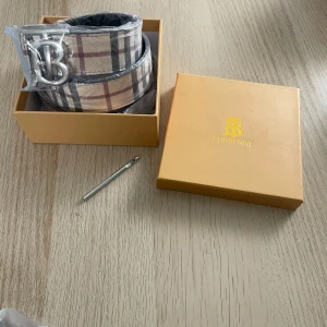Burberry bälte med silverfärgat spänne - Säljer ett snyggt bälte från Burberry med klassiskt rutigt mönster i beige, svart och rött. Bältet har ett stort silverfärgat B-spänne och är vändbart med svart mönstrad insida. Perfekt accessoar för att lyfta din outfit. För mer info hör av dig. St 115